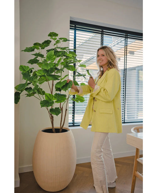 Una mujer con una chaqueta amarilla y pantalones blancos sonríe mientras cuida la Planta Artificial Ficus de 150 cm en una habitación moderna y bien iluminada, con suelos de madera y grandes ventanales con persianas.