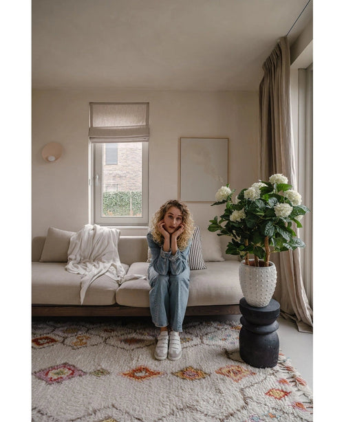 Una mujer con cabello rubio y rizado se sienta en un sofá beige en una sala de estar luminosa y apoya la barbilla en las manos. Junto a ella se encuentra la planta UV Hortensia Artificial de 85 cm de altura, iluminada por la luz a través de una ventana con cortinas beige.