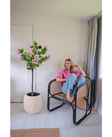 Una mujer con un jersey rosa y vaqueros azules está sentada descalza en una silla de ratán junto a la Planta Artificial Judas 150 cm, un árbol artificial con flores rosas, en una habitación moderna y luminosa con paredes y suelos blancos.