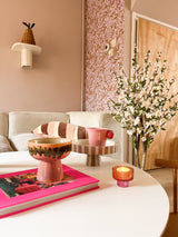 En un acogedor salón hay una mesa de centro blanca con un libro rosa, un cuenco, un cuenco con pedestal, una vela y la planta de cerezo artificial color crema de 120 cm. Al fondo hay un sofá beige con cojines a rayas y papel pintado con motivos rosas.