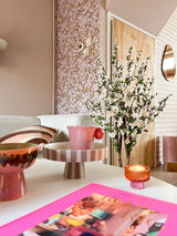 Una acogedora sala de estar con una decoración rosa y pastel presenta una bandeja a rayas, una taza rosa, una vela, un cuenco, una revista animada y la planta de cerezo artificial color crema de 120 cm en un jarrón alto cerca de una pared estampada y una puerta de madera.