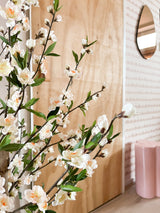 Delante de una puerta de madera hay una planta de cerezo artificial de 120 cm color crema con flores blancas y rosa claro. Detrás hay un taburete rosa, un espejo y papel pintado con motivos rosas.