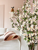 Una acogedora sala de estar con un sofá blanco, un cojín a rayas rosas y marrones y en primer plano la rama floreciente de cerezo artificial color crema de 120 cm. Una planta en maceta descansa sobre un estante de pared contra una pared de color rosa pálido.