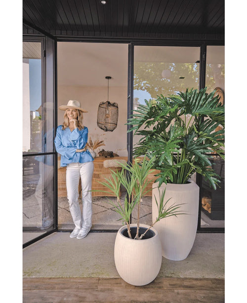 Una mujer con sombrero, blusa azul y pantalón blanco se apoya contra una puerta de cristal. En primer plano se encuentra la planta artificial Filodendro de 125 cm. En el interior se puede ver un espacio habitable acogedor y moderno.