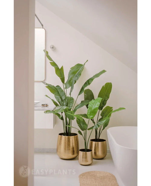 Una planta artificial Strelitzia Nicolai Deluxe de 120 cm se encuentra en una maceta dorada brillante en el suelo de un baño moderno y minimalista con bañera blanca y paredes de colores claros.