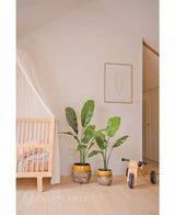 Una acogedora habitación infantil con una cuna de madera, una lujosa planta artificial Strelitzia Nicolai de 120 cm, un dibujo lineal minimalista enmarcado en la pared y una pequeña bicicleta sin pedales de madera con la etiqueta MIA sobre un suelo de madera clara.