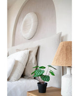 Una cama cuidadosamente hecha con almohadas y ropa de cama blancas se encuentra junto a una mesita de noche de madera con la planta de Sandía Artificial de 29 cm y una lámpara con pantalla tejida; encima de la cabecera cuelga una escultura de pared con textura blanca.