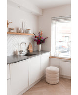 Una cocina moderna y minimalista con armarios blancos, una encimera negra y una flor de hortensia artificial de 55 cm de color rosa oscuro en un jarrón cerca de la encimera, además de estantes de madera con decoración y un taburete redondo con cojines cerca de una ventana con persianas.