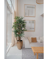 Una Lady Artificial Palm de 180 cm se coloca cerca de un gran ventanal en una sala de estar moderna y luminosa con un sofá blanco, cojines beige, una mesa tejida y arte abstracto. Agrega un estilo minimalista acogedor con sus tonos neutros y el encanto de las plantas artificiales verdes.