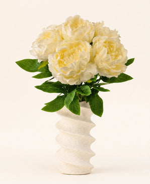 En un jarrón blanco ondulado y estructurado se encuentra el ramo de flores artificiales de seda Peony White: grandes peonías artificiales de color crema con hojas verdes sobre un fondo liso y claro.