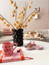 Un acogedor salón con un sofá beige, un gato durmiendo, un escultural jarrón negro que contiene la Flor Artificial Physalis Lampion marrón de 90 cm, una taza rosa "LOVE", un libro, una vela y una galleta sobre la mesa de centro.
