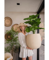 Una mujer con un vestido blanco y un sombrero sonríe mientras sostiene la planta Artificial Calathea UV de 72 cm en el interior, con cestas tejidas y otra planta al fondo.
