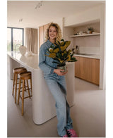 Una persona sonriente con ropa vaquera se encuentra en una cocina moderna sosteniendo una planta de Magnolia artificial en una maceta de 46 cm, una planta de fácil cuidado. La cocina, perfecta para la decoración de interiores, cuenta con gabinetes de madera, una isla de cocina blanca y dos taburetes de bar.