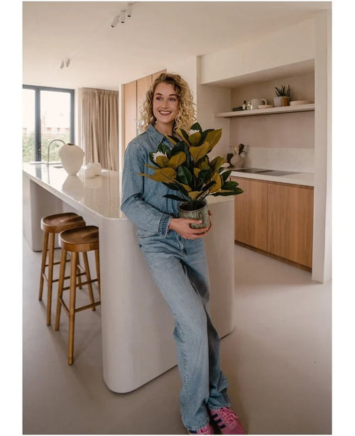 Una persona sonriente con ropa vaquera se encuentra en una cocina moderna sosteniendo una planta de Magnolia artificial en una maceta de 46 cm, una planta de fácil cuidado. La cocina, perfecta para la decoración de interiores, cuenta con gabinetes de madera, una isla de cocina blanca y dos taburetes de bar.