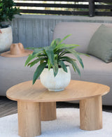 Una planta artificial Bird's Deluxe de 60 cm se encuentra sobre una mesa de centro redonda de madera frente a un sofá beige, decorada con un cojín verde y un sombrero marrón que cuelga sobre el reposabrazos.