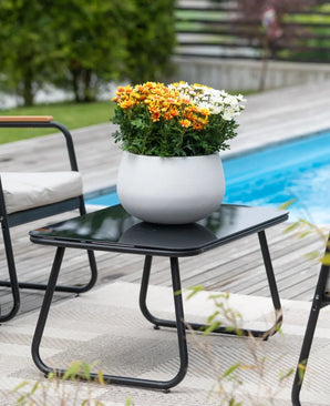 Una mesa de patio negra con una planta Artificial Daisy UV de 31 cm (una maceta blanca redonda con vibrantes margaritas de color naranja y blanco) se encuentra junto a una piscina, rodeada de sillas de jardín y con vegetación de fondo.