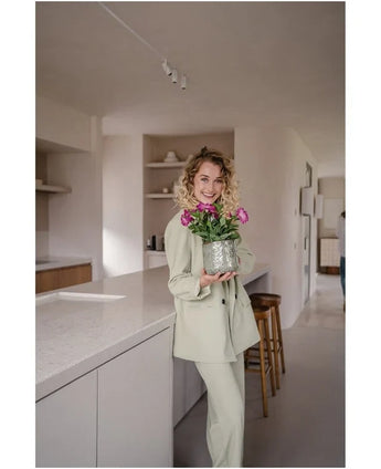 Una mujer con un traje claro sonríe mientras sostiene una Flor Artificial Petunia UV 25 cm rosa oscuro; ella está parada en una cocina moderna con tonos neutros y decoración minimalista.