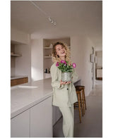 Una mujer sonriente, de pelo rizado y vestida con un traje de color claro, se encuentra en una cocina moderna y minimalista sosteniendo una flor artificial Petunia UV de 25 cm de color rosa oscuro con flores de color rosa intenso.