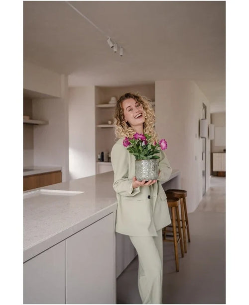 Una mujer sonriente, de pelo rizado y vestida con un traje de color claro, se encuentra en una cocina moderna y minimalista sosteniendo una flor artificial Petunia UV de 25 cm de color rosa oscuro con flores de color rosa intenso.