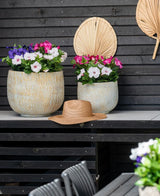 Dos grandes maceteros de cerámica con una Flor Artificial Petunia UV de 25 cm de color rosa oscuro, con flores de un rosa vibrante, se encuentran sobre una mesa contra una pared de madera oscura. Hay un sombrero de paja sobre la mesa y hojas de palma secas cuelgan de la pared encima de las flores.