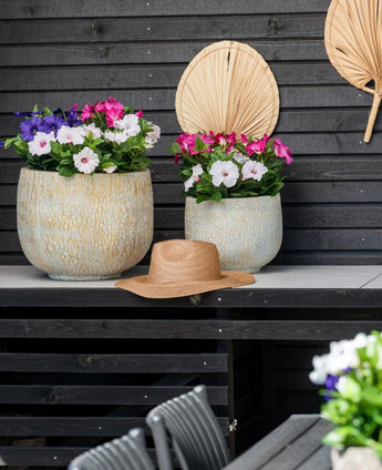 Dos grandes maceteros de cerámica con una Flor Artificial Petunia UV de 25 cm de color rosa oscuro, con flores de un rosa vibrante, se encuentran sobre una mesa contra una pared de madera oscura. Hay un sombrero de paja sobre la mesa y hojas de palma secas cuelgan de la pared encima de las flores.