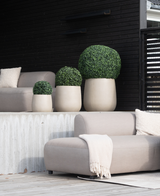 Vier Kunstplant Buxus romeinse stijl UV D40cm, rond en beige, met verzorgde groene struiken, staan op een moderne patio met lichtgrijze stoelen, een beige deken en een donkerhouten achtergrond voor een minimalistische look.