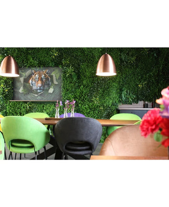 Een moderne eethoek met groene en perzikkleurige stoelen, een tafel versierd met vazen met paarse bloemen, koperen hanglampen, een weelderige plantenwand gemaakt met het Kunsthaag Crush UV-paneel van 100 x 100 cm, en op de achtergrond een ingelijste afbeelding van een tijger.