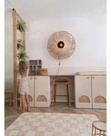 Een jong meisje in een lichte jurk staat naast een plank waarop de Kunst-hangplant ‘Frosted Varen op bal 76 cm’ en andere hangplanten staan, in een moderne kamer met beige kasten, een hoge kruk, een wandlamp van riet en een geruit vloerkleed op de vloer.