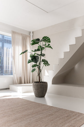 La Planta Artificial Monstera 210 cm se encuentra en una maceta sobre un suelo blanco junto a una escalera moderna en una habitación luminosa y minimalista con luz natural que entra a través de cortinas beige.