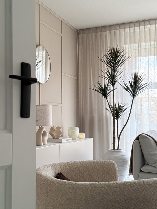 Un salón moderno y minimalista con un sillón con textura beige, una planta artificial Dracaena Fragnans de 180 cm, un aparador blanco con decoración, un espejo redondo y cortinas transparentes que filtran la suave luz del día a través de la ventana.