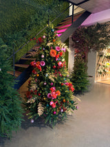 El árbol de Navidad artificial "Weideman" PE/PVC - Smart LED se encuentra en el interior cerca de una escalera, decorado con flores rosas y rojas, hojas doradas y adornos rosas, rodeado de vegetación sobre un suelo de hormigón pulido para una elegante decoración navideña.
