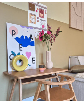 Un pequeño escritorio de madera con una silla está decorado con una flor artificial de hierba gatera rosa de 75 cm, una taza, una escultura de un anillo amarillo y coloridas impresiones artísticas en la pared de arriba. A la derecha se ve parcialmente un banco.