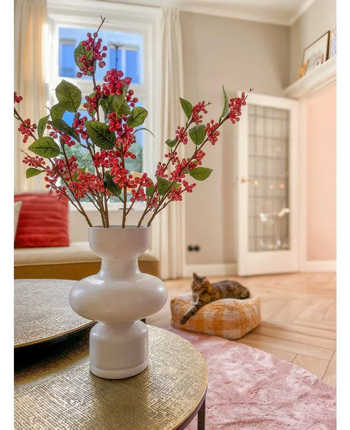Sobre una mesa hay un jarrón blanco con bayas rosas de Kunsttak (64 cm) y hojas verdes. Al fondo, un gato tumbado sobre un cojín rosa en un luminoso salón con paredes beige y una puerta de cristal.