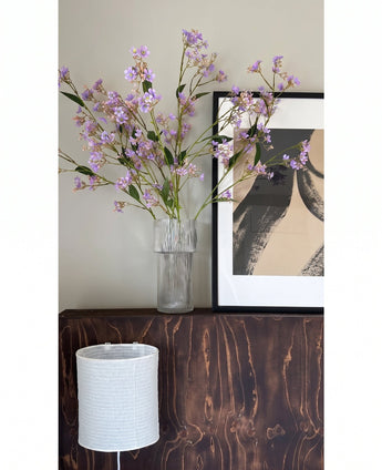 En un jarrón transparente, las flores del Kunsttak Daphne Spray 108 cm violeta se colocan sobre una superficie de madera oscura junto a una obra de arte moderna y abstracta enmarcada, con una pantalla estructurada blanca debajo.
