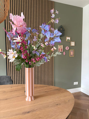 Un jarrón alto a rayas rosas y blancas con el ramo de flores artificiales de seda Amethyst Romance se encuentra sobre una mesa redonda de madera en una habitación moderna con paredes verdes, paneles de madera y pequeños cuadros enmarcados.