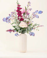 Un jarrón blanco texturizado exhibe el "Ramo de flores artificiales de seda Amethyst Romance" con peonías rosadas, flores violetas y azules, flores color burdeos y follaje verde sobre un fondo claro y neutro.