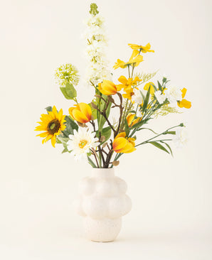 In een witte vaas met structuur staat het Zijden kunstbloemen boeket Sun Glow met gele tulpen, zonnebloemen, madeliefjes en groen blad tegen een effen gebroken witte achtergrond.