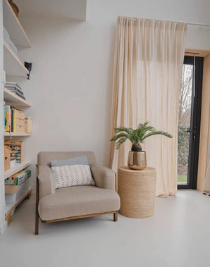 Un acogedor rincón de lectura con un sillón beige, una mesa auxiliar redonda de mimbre con la planta Baby Cycas artificial de 60 cm, estanterías y visillos que cuelgan sobre una puerta de cristal que da al jardín.