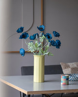 Una flor de amapola artificial de 73 cm en color azul oscuro se encuentra en un jarrón amarillo sobre una mesa, acompañada de una pequeña taza a rayas de colores, en una habitación moderna, suavemente iluminada, con una decoración minimalista y tonos neutros.