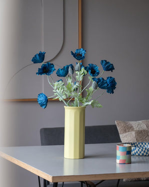 Una flor de amapola artificial de 73 cm en color azul oscuro se encuentra en un jarrón amarillo sobre una mesa, acompañada de una pequeña taza a rayas de colores, en una habitación moderna, suavemente iluminada, con una decoración minimalista y tonos neutros.