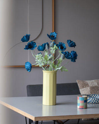 Una flor de amapola artificial de 73 cm en color azul oscuro se encuentra en un jarrón amarillo sobre una mesa, acompañada de una pequeña taza a rayas de colores, en una habitación moderna, suavemente iluminada, con una decoración minimalista y tonos neutros.