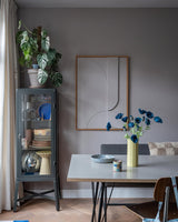 La moderna zona de comedor tiene una vitrina de cristal gris en la que se exponen vajillas y plantas, decoraciones minimalistas en las paredes y una mesa sobre la que se encuentra la flor artificial de amapola de 73 cm de color azul oscuro en un jarrón amarillo. El espacio se completa con una mezcla ecléctica de sillas, todo ello sobre un fondo de paredes grises y suelo de madera.