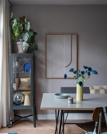 La moderna zona de comedor tiene una vitrina de cristal gris en la que se exponen vajillas y plantas, decoraciones minimalistas en las paredes y una mesa sobre la que se encuentra la flor artificial de amapola de 73 cm de color azul oscuro en un jarrón amarillo. El espacio se completa con una mezcla ecléctica de sillas, todo ello sobre un fondo de paredes grises y suelo de madera.