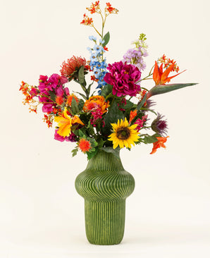 Un jarrón estructurado de color verde muestra el ramo de flores artificiales de seda Colorful Spirit, con girasoles, peonías rosas, flores de color naranja y violeta y exuberante vegetación sobre un fondo claro y sencillo.