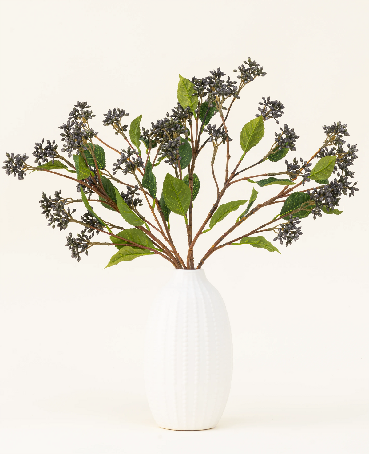 Un ramo de flores artificiales de seda de bayas japonesas de color azul acero con tallos largos, bayas de color púrpura oscuro y hojas de acero se encuentra en un jarrón con textura blanca sobre un fondo claro y liso.