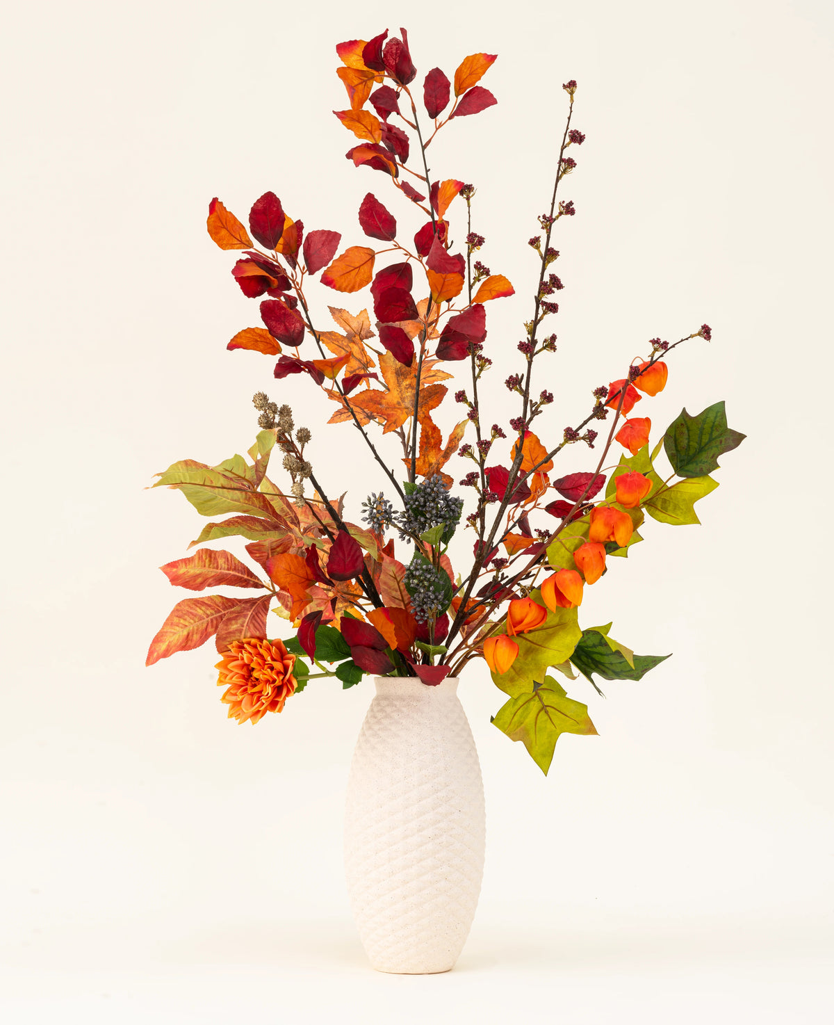 Un jarrón con textura blanca exhibe el ramo de flores artificiales de seda con dijes de castaño, que presenta hojas de otoño, flores de color naranja y follaje verde: un elegante arreglo artificial sobre un fondo liso y claro.