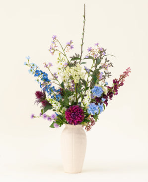 Un jarrón blanco con el ramo de flores artificiales de seda Purple Rain muestra vibrantes flores artificiales de color púrpura y blanco y hojas verdes, un ejemplo de artesanía holandesa sobre un fondo claro.