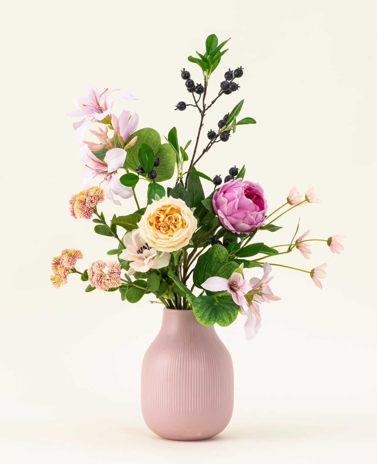 El ramo de flores artificiales de seda Touch of Cloudlight, presentado en un jarrón de cerámica rosa, contiene peonías rosadas, rosas amarillas, pequeñas flores, vegetación y ramas con bayas oscuras sobre un fondo claro.