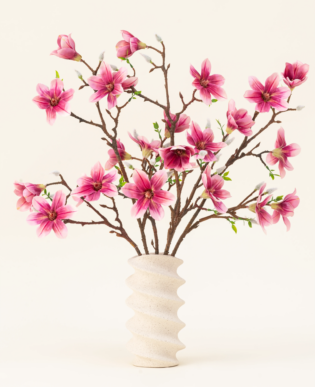 El ramo de flores artificiales de seda Magnolia rosa está dispuesto en un jarrón de cerámica blanco ondulado con capullos verdes, creando una elegante exhibición sobre un fondo claro.