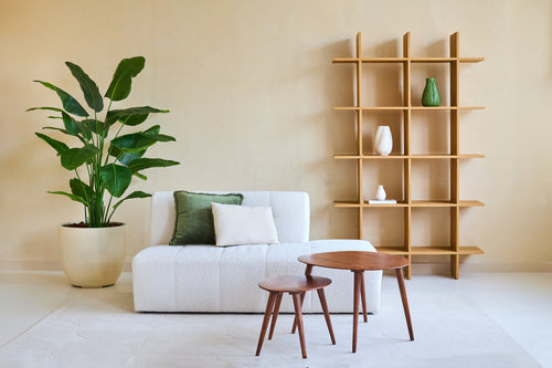 Salón minimalista con sofá blanco, dos cojines, mesas nido de madera y una gran Planta Artificial Strelitzia Real Touch Deluxe 180 cm para un ambiente tropical. Contra la pared beige hay una moderna estantería abierta de madera con jarrones.
